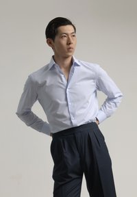 Camisa de vestir de manga larga en azul claro con un patrón a cuadros y collar abotonado, combinada con pantalones oscuros con pliegues, de pie sobre un fondo neutral.
