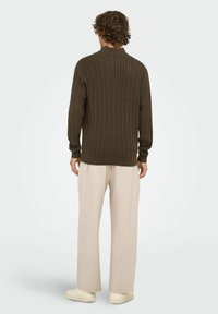 Hombre con cabello rizado, vistiendo un suéter de punto trenzado marrón oscuro, pantalones anchos beige y zapatillas de color claro, de pie de espaldas.