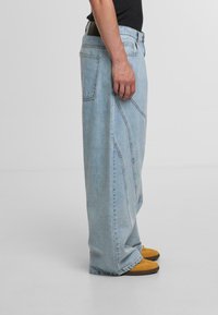 Helle blaue Denim-Jeans im weiten Schnitt mit sichtbaren Nähten und hoher Taille, versehen mit einem schwarzen Gürtel. Getragen mit braunen und gelben Schuhen.
