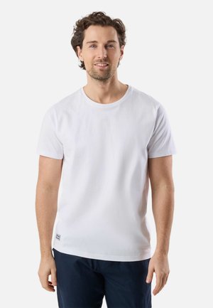 Mand med krøllet brunt hår iført en ensfarvet hvid kortærmet t-shirt og mørkeblå bukser mod en hvid baggrund.