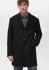 Abrigo de mezcla de lana negro con cuello de muesca, diseño de un solo botón, cuatro botones y bolsillos laterales. Llevado sobre una camisa de botones gris.