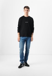 Jonge man die staat in een eenvoudige studio-instelling, gekleed in een zwart langemouwen shirt, blauwe spijkerbroeken en zwarte sneakers, met een neutrale uitdrukking.