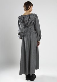 Mujer de espaldas vistiendo un vestido a cuadros en blanco y negro, con mangas abullonadas y zapatos de plataforma negros, sobre un fondo liso.