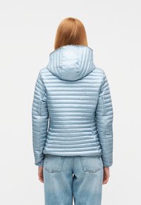 Γυναίκα με μακριά κόκκινα μαλλιά, που φορά ελαφρύ μπλε υφασμάτινο μπουφάν με κουκούλα και μπλε τζιν, εικονιζόμενη από πίσω πάνω σε λευκό φόντο.