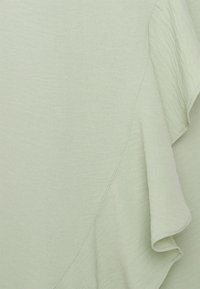 Vêtement vert clair en tissu doux et texturé. Présente un détail de volants fluides le long du bord et une couture subtile en diagonale.