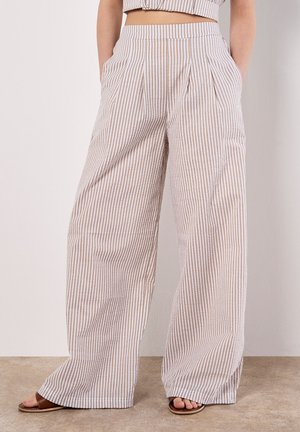Beżowe i białe spodnie w pionowe paski o szerokich nogawkach, noszone z brązowymi sandałami na odkryte palce, modelka stoi z rękami w kieszeniach.