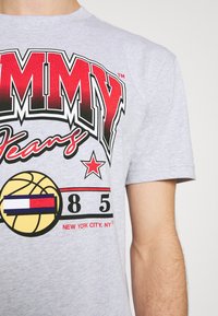 Vaaleanharmaa T-paita, jossa on iso punainen ja musta "Tommy" -teksti, koripallografiikka, punainen tähti ja "New York City, NY" painettuna eteen.