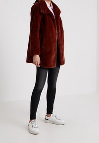 Brązowy płaszcz z sztucznego futra z dużym kołnierzem, o luźnym kroju i długich rękawach, zestawiony z czarnymi jeansami i białymi tenisówkami.