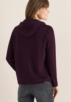 Sudadera morada con un bolsillo canguro en la parte delantera, mangas largas y una textura suave. Combinada con jeans grises que tienen bolsillos traseros.