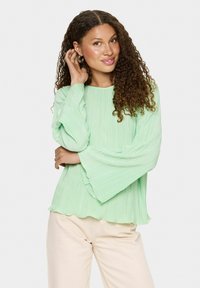 Jasnozielona, plisowana bluzka z dzwonowymi rękawami i falbaniastym dołem; noszona z kremowymi spodniami. Subtelny połysk i zwiewna faktura.