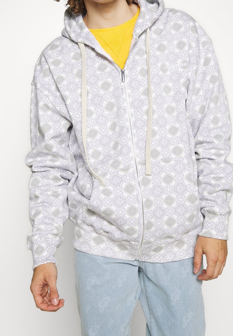 Personne portant un sweat à capuche zippé gris clair à motifs sur un t-shirt jaune et un jean en denim bleu clair, debout les bras détendus.