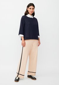 Maglione navy con maniche larghe sopra una camicia bianca con colletto, abbinato a pantaloni larghi color pesca chiaro con righe laterali nere e sneakers nere.