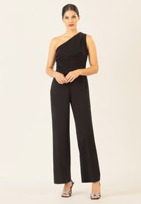 Jumpsuit nero a spalla unica con tessuto drappeggiato, gambe ampie e una texture liscia. Abbinato a tacchi neri con un fiocco decorativo.