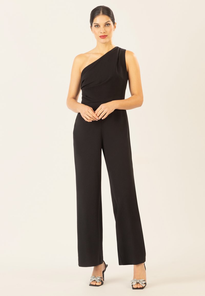 Jumpsuit nero a spalla unica con tessuto drappeggiato, gambe ampie e una texture liscia. Abbinato a tacchi neri con un fiocco decorativo.