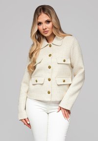 Kremowa krótka marynarka z tweedu z dużymi złotymi guzikami, dwoma kieszeniami na piersi i spiczastym kołnierzem, zestawiona z białymi spodniami.