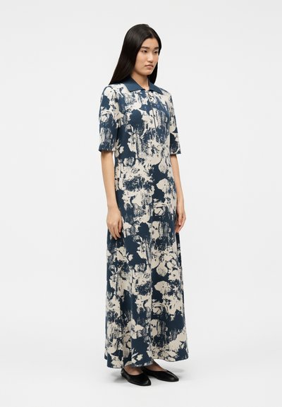 Γυναίκα στέκεται φορώντας μακρύ φόρεμα με αφηρημένα λουλούδια σε ναυτικό μπλε και λευκό χρώμα, με κοντά μανίκια, γιακά και μαύρα επίπεδα παπούτσια.