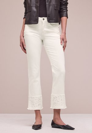 Kobieta ubrana w białe spodnie 7/8 z koronkowym brzegiem z oczkami, czarne spiczaste baleriny i czarną skórzaną kurtkę, stojąca na beżowym tle.