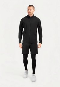 Czarna sportowa kurtka z pełnym zamkiem błyskawicznym, w połączeniu z czarnymi szortami i legginsami. Szare sneakersy. Prosty design i gładka faktura.