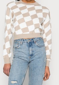 Suéter corto beige y blanco con un patrón de diamantes, puños acanalados y dobladillo, combinado con jeans rasgados de color azul claro.
