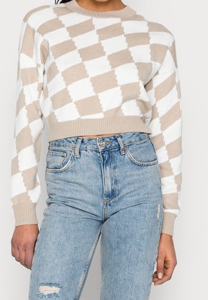 Suéter corto beige y blanco con un patrón de diamantes, puños acanalados y dobladillo, combinado con jeans rasgados de color azul claro.