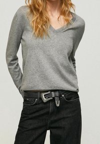 Pull gris à col en V, manches longues, texture douce. Associé à un jean foncé et une ceinture noire avec une boucle argentée et des coutures détaillées.