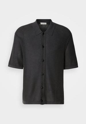 Chemise noire à manches courtes avec une texture en maille, devant à boutons et col. Présente un ourlet lisse et un logo discret en bas.