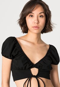 Top cropped preto com mangas bufantes, decote profundo e detalhe central em forma de buraco. Tecido suave e elástico com costuras franzidas. Atado nas costas.