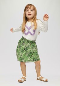 Weißes Langarmhemd mit lila strukturiertem Muster, kombiniert mit einem grünen Blümchenrock und weißen Sandalen. Einfaches, lässiges Kinderoutfit.