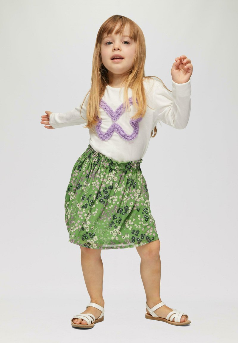 Weißes Langarmhemd mit lila strukturiertem Muster, kombiniert mit einem grünen Blümchenrock und weißen Sandalen. Einfaches, lässiges Kinderoutfit.