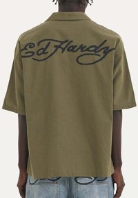 Olijfgroene shirt met een losse pasvorm, korte mouwen en een groot zwart "Ed Hardy" logo geborduurd op de achterkant. Zacht, textuurstof.