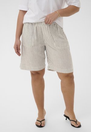 Person iført beige og sorte shorts med lodrette striber, frontlommer, en hvid skjorte, guldbøjler og sorte sandaler med hæl.