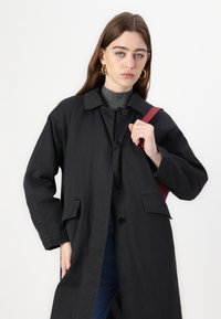 Black cotton long coat with a notched collar, four buttons, and front pockets, worn over a gray top. The model holds a red shoulder strap.