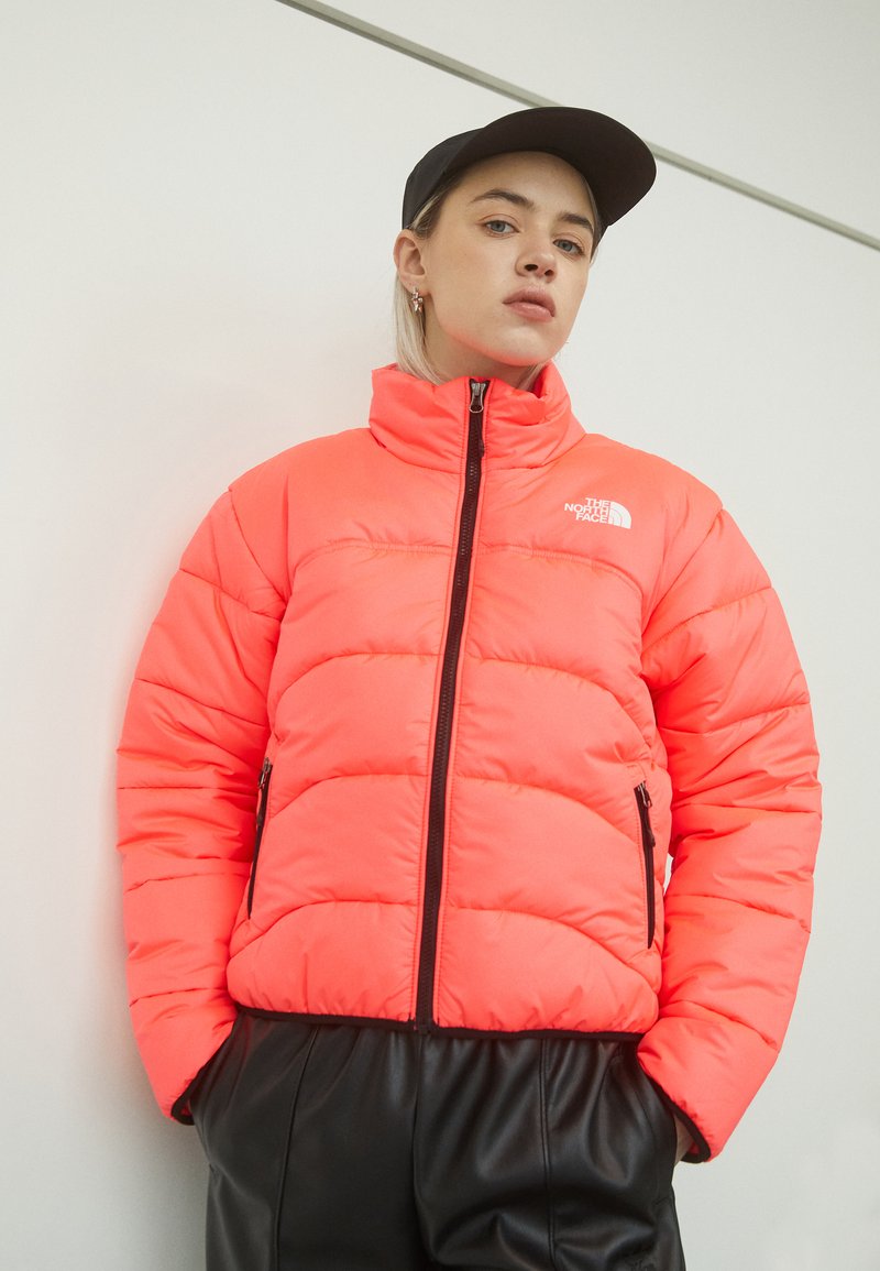 Giacca imbottita neon rosa brillante con colletto alto, chiusura frontale con zip e dettagli neri. Presenta due tasche laterali con zip e cuciture orizzontali.