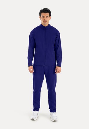 Hombre de pie con chaqueta azul oscuro con cremallera y pantalones a juego, con zapatillas blancas, sobre un fondo liso y claro.