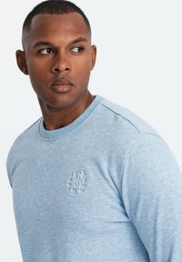 Sudadera de algodón azul claro con cuello redondo, que presenta un logo circular texturizado en el lado izquierdo del pecho y puños acanalados.