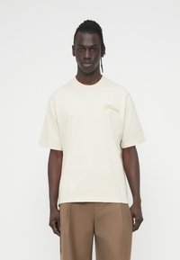 Μπεζ βαμ βακερό t-shirt με κοντά μανίκια, στρογγυλή λαιμόκοψη και διακριτικό λογότυπο κεντημένο σε ανοιχτότερη απόχρωση στην αριστερή πλευρά.