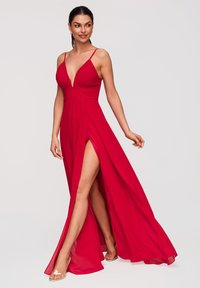 Femme portant une robe rouge fluide à fines bretelles, avec un profond décolleté en V et une fente haute à la cuisse, posant avec une jambe dégagée et des talons transparents.