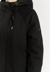 Sort hættejakke lavet af tekstureret stof med en frontzipper, snoretræk og en fremtrædende logo-nål på ærmet.