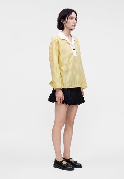 Femme debout portant une chemise jaune à manches longues avec col blanc, une jupe courte noire avec ourlet à volants et des chaussures Mary Jane noires.