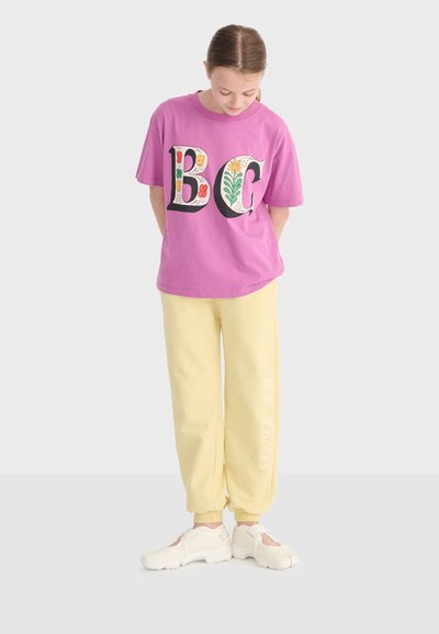 Pige iført en lilla T-shirt med blomstrede "BG"-bogstaver, blege gule joggingbukser og hvide sko, der kigger ned med hænderne bag ryggen.