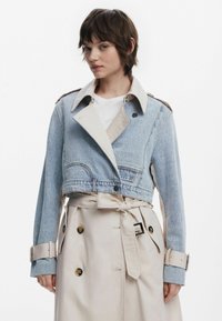 La chaqueta recortada presenta un denim de color azul claro con un cuello beige en contraste, grandes botones y detalles de cinturón en la cintura y las mangas.