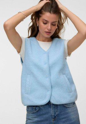 Femme ajustant ses cheveux, portant une veste polaire bleu clair sur un t-shirt blanc à manches courtes et un jean bleu.