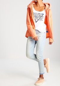 Chaqueta impermeable naranja claro con capucha con patrón, camiseta de tirantes blanca con gráfico, vaqueros ajustados azul claro y zapatillas beige.