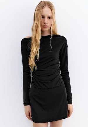 Jeune femme aux longs cheveux blonds portant une robe noire ajustée à épaules dénudées, debout devant un fond blanc uni.