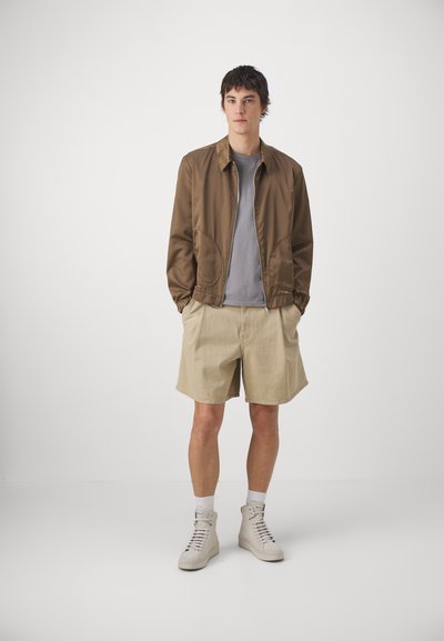 Giacca bomber marrone sopra una maglietta grigia, abbinata a pantaloni corti beige e scarpe da ginnastica leggere. Presenta una chiusura con zip e tasche laterali.