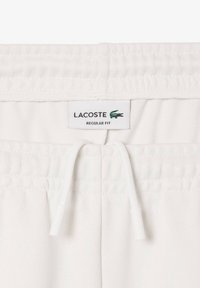 Pantalon blanc à cordon de serrage avec taille élastique, tissu texturé et étiquette "Lacoste REGULAR FIT" avec logo crocodile vert.