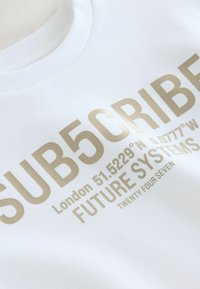 Vit sweatshirt med guldtext: "SUB5CRIBE", koordinater och "FUTURE SYSTEMS" med ribbad halsringning och mjuk textur.