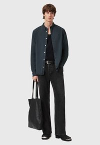 Camicia button-up blu scuro sopra una canottiera nera, abbinata a jeans neri e una borsa a tracolla nera con texture, che presenta un logo discreto sulla camicia.