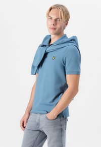 Blå bomullspolo med en gul logo, parret med lysegrå jeans. En blå genser er nonchalant drapert over skuldrene.