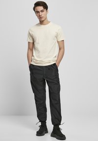 Lichtbeige katoenen t-shirt gecombineerd met zwarte, lichte nylon broeken met een relaxte pasvorm en elastische boorden. Zwarte sneakers maken de outfit af.